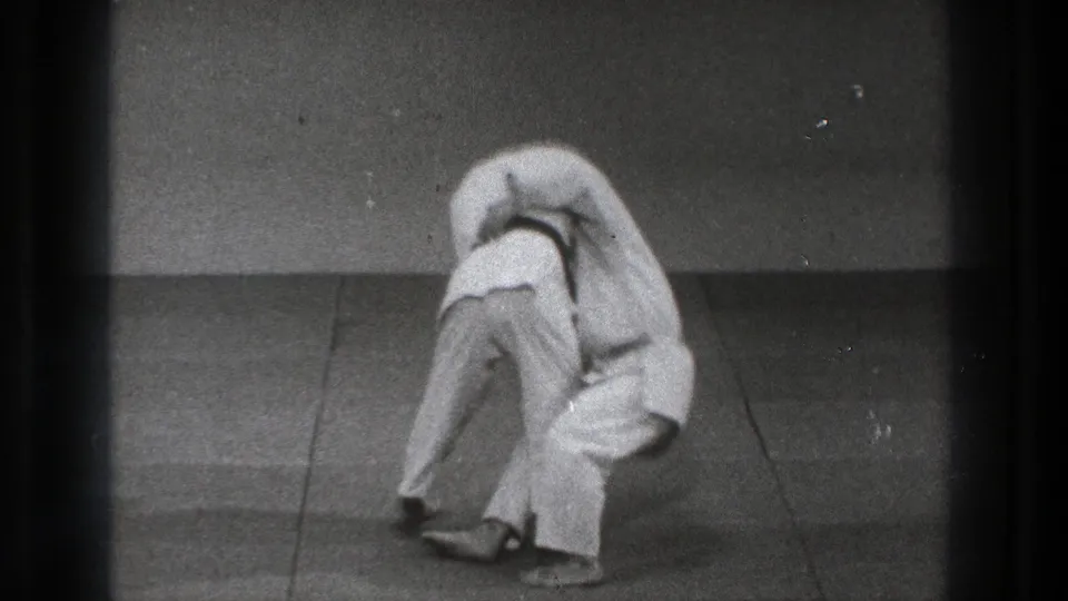 The Tokyo martial arts sequence shows one adult in a white gi lifting another into the early phase of a throw.