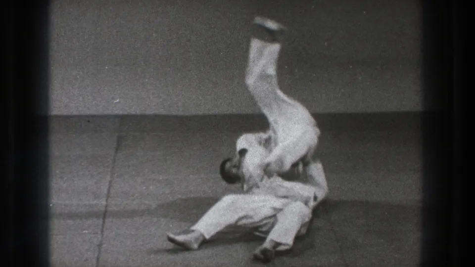 The throw reaches its peak as one adult in a white gi flips upside down over another practitioner on the Tokyo training mat.