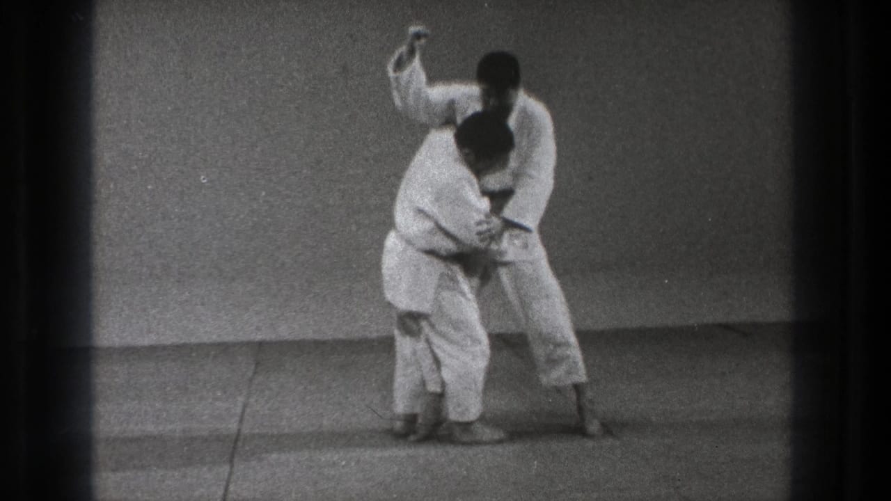 Two martial artists in white gi grapple on a mat as one raises an arm.