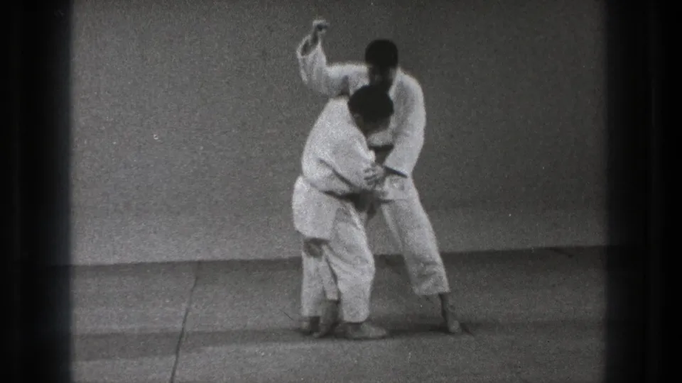 In metadata-supported Tokyo, 1971, two martial artists in white gi begin a judo-style hold on a plain indoor mat.