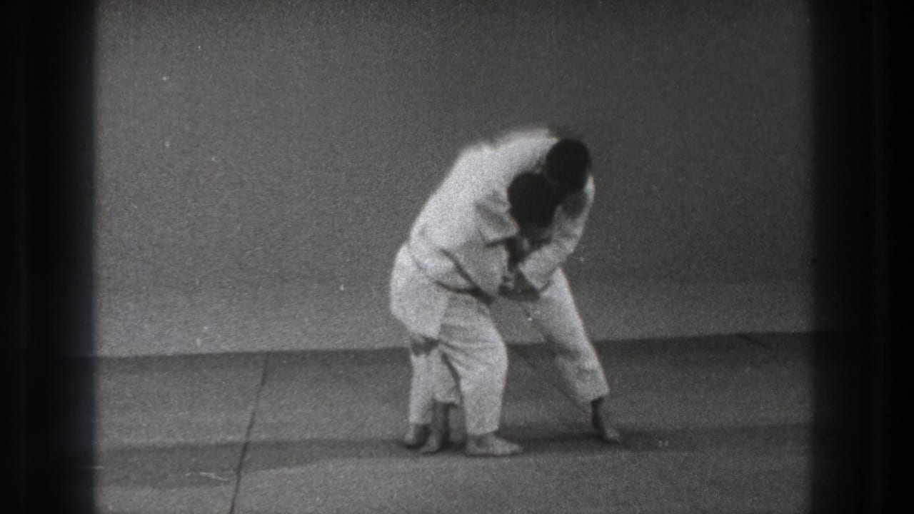 A martial artist in white gi leans into the throw setup while the second practitioner stays close at hip level.
