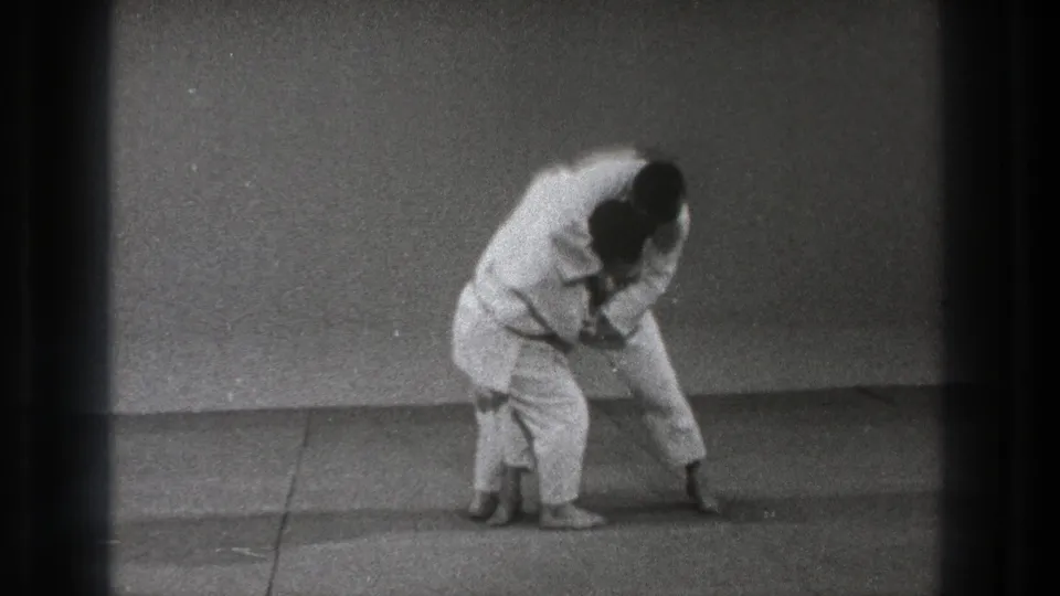 A martial artist in white gi leans into the throw setup while the second practitioner stays close at hip level.