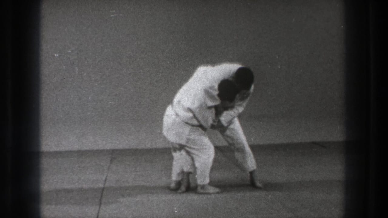 Two martial artists in white gi bend together on a mat during a defensive technique.