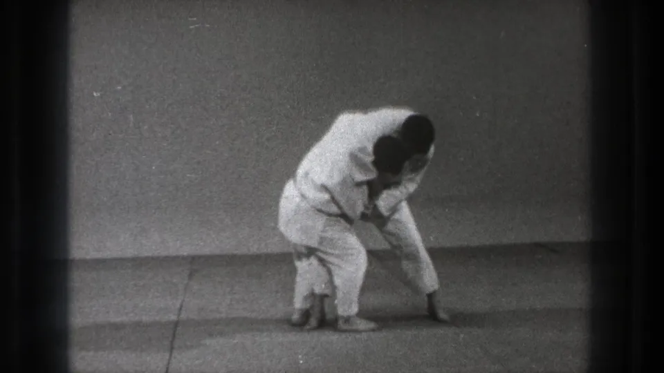In 1971 Tokyo footage, two martial artists in white gi lean into a controlled defensive movement on a plain training mat.