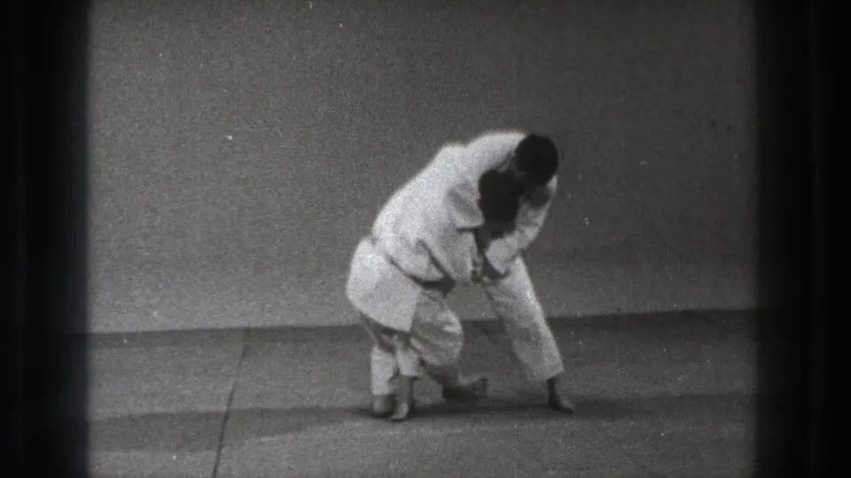 Two karate practitioners in white gi demonstrate low stance and partner control against a plain studio backdrop in Tokyo-era footage.