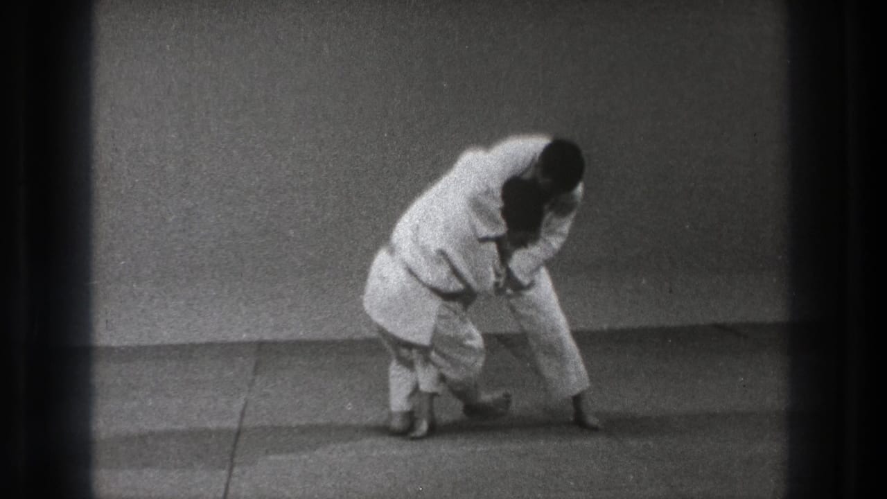 A white-gi practitioner bends over a partner during a controlled karate defense demonstration on a simple mat surface.