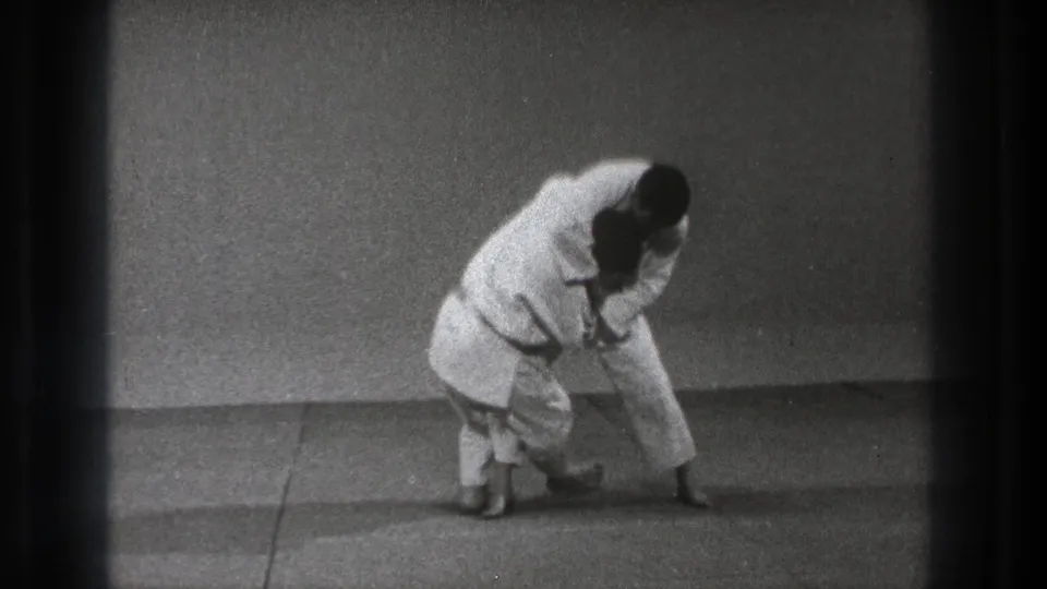 A white-gi practitioner bends over a partner during a controlled karate defense demonstration on a simple mat surface.