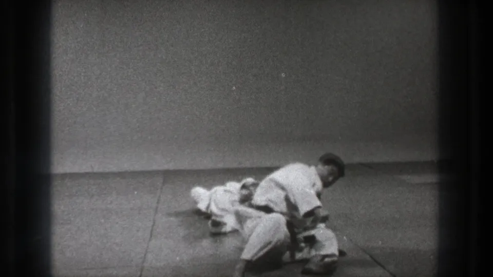 1971 Tokyo home-movie frame showing two adult men in white uniforms practicing a ground maneuver on indoor mats.