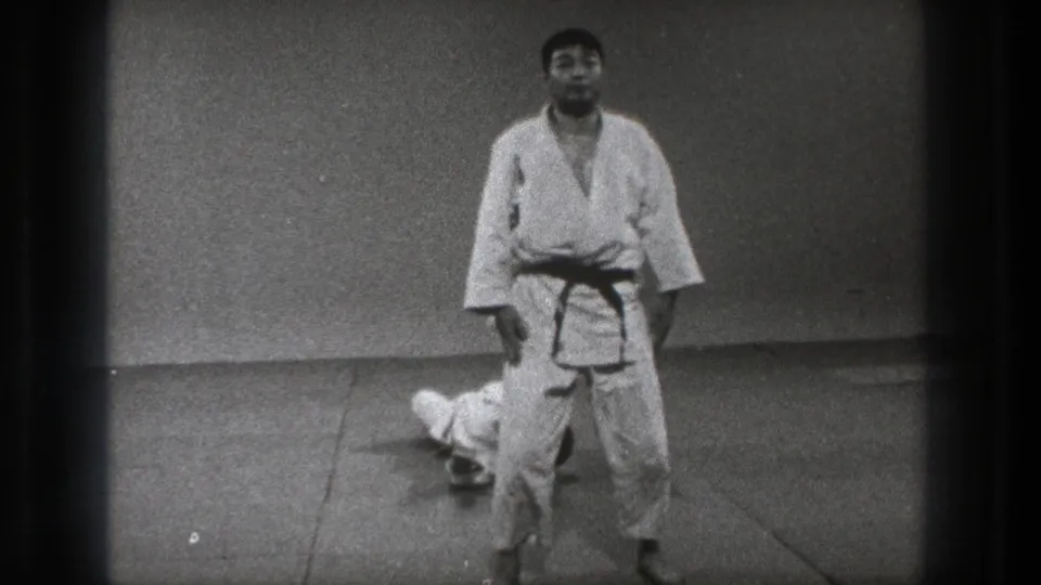 1971 Tokyo frame with one martial arts practitioner standing in a formal posture as another remains on the indoor mat.