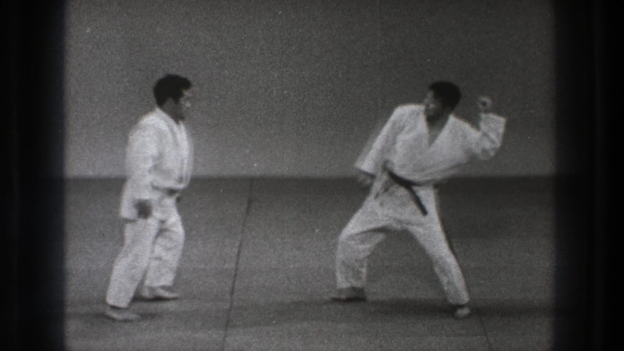 1971 Tokyo home-movie frame showing two adult practitioners preparing for a martial arts maneuver on mats.