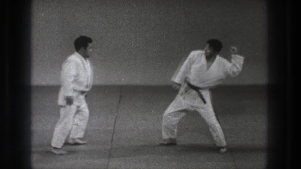1971 Tokyo home-movie frame showing two adult practitioners preparing for a martial arts maneuver on mats.