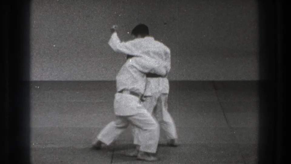 In 1971 Tokyo, two martial artists in white gi begin a close judo throw practice sequence on a plain indoor mat.