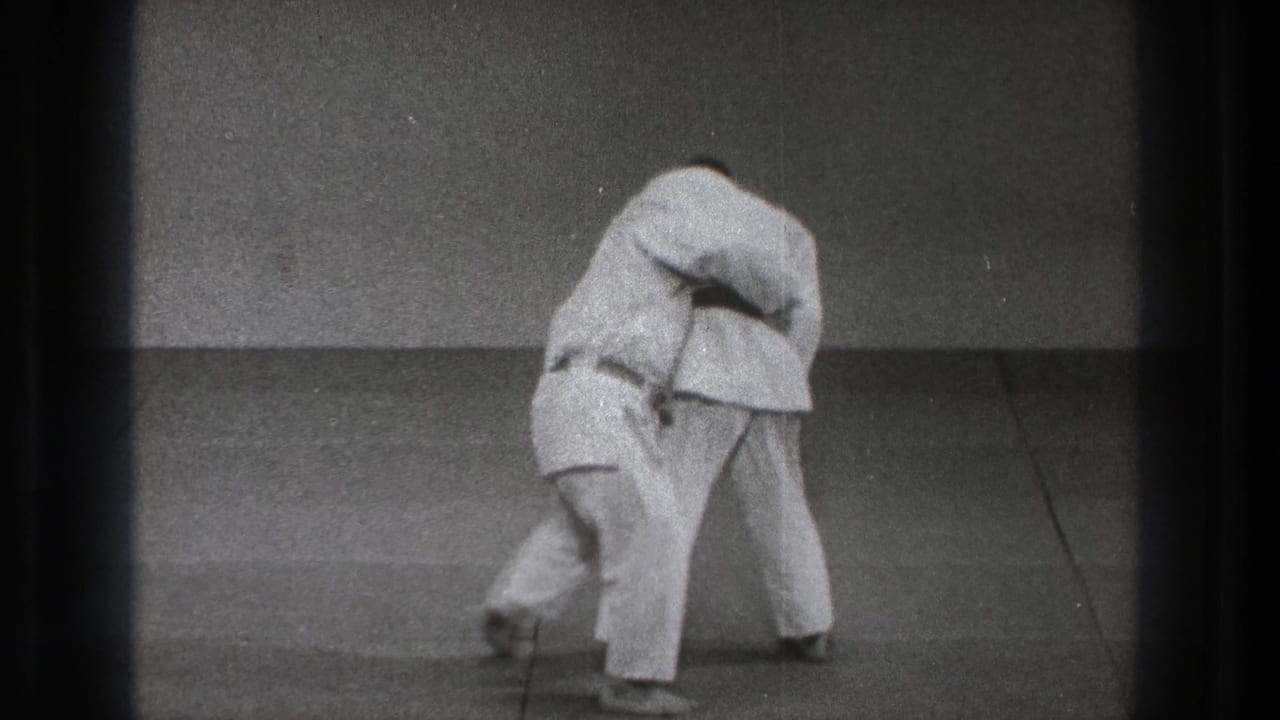 The paired judo practice reaches the throwing motion, with one martial artist bent low beneath the other's grip on the mat.