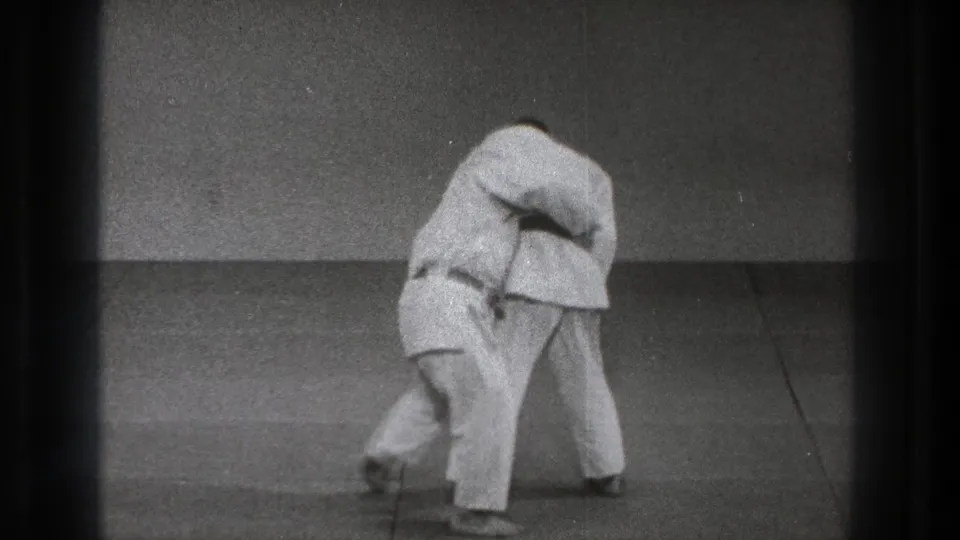 The paired judo practice reaches the throwing motion, with one martial artist bent low beneath the other's grip on the mat.