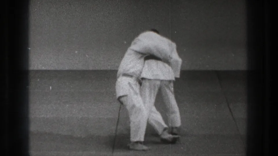 Near the end of the 1971 Tokyo judo sequence, one practitioner drops low as the other controls the grip during the throw practice.