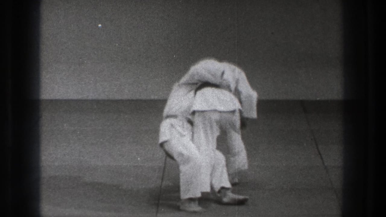 Two martial artists in white gi begin a throw on a plain mat.