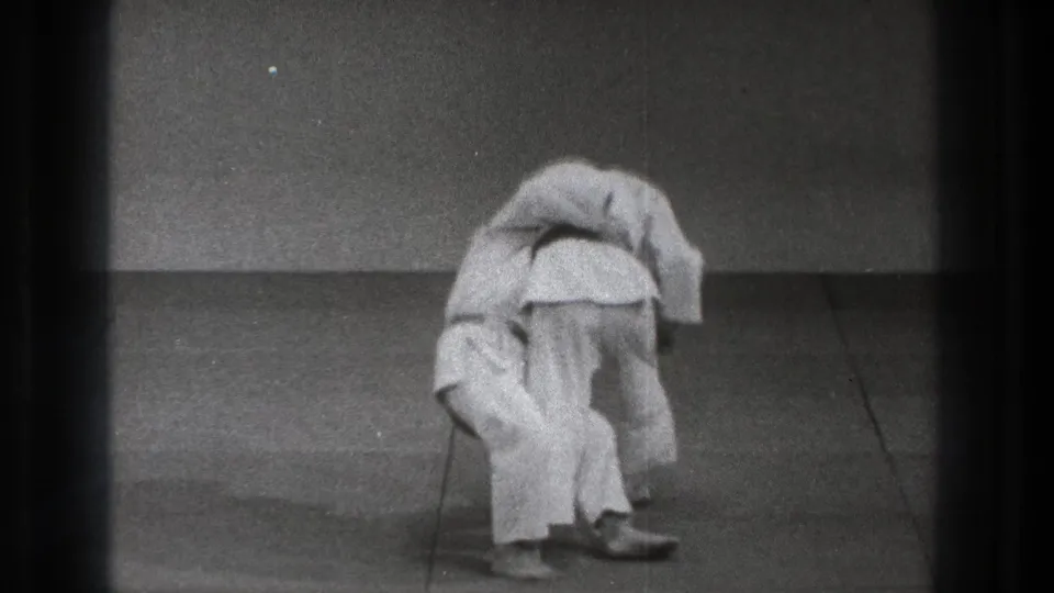 In supplied 1971 Tokyo footage, one martial artist in a white gi bends forward while controlling another during a throw setup on a plain mat.