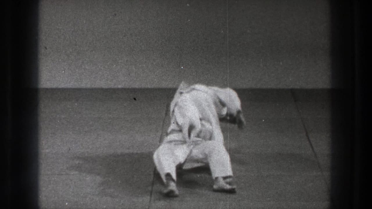 A gi-clad practitioner leans and falls backward while the partner remains close, continuing the takedown in the sparse indoor practice space.