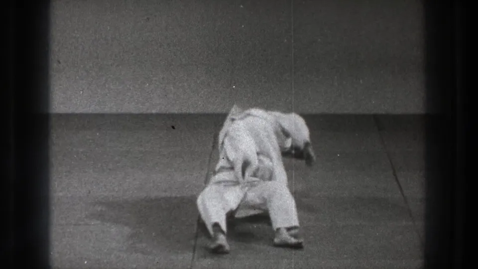 A gi-clad practitioner leans and falls backward while the partner remains close, continuing the takedown in the sparse indoor practice space.