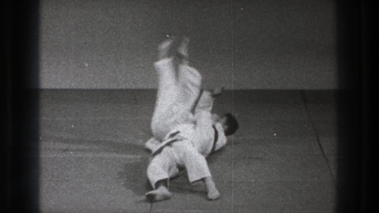 During the throw sequence, one practitioner is inverted above the other as both white gi contrast against the dark padded floor.