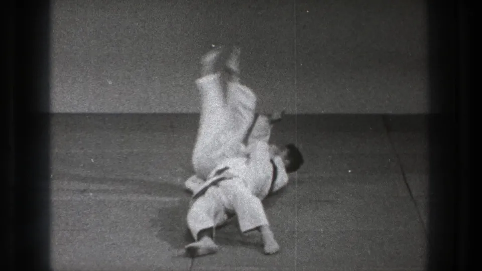 During the throw sequence, one practitioner is inverted above the other as both white gi contrast against the dark padded floor.