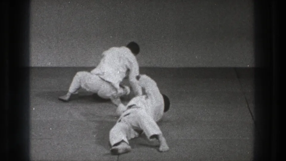 In 1971 Tokyo footage, two adult men in white martial arts uniforms practice a ground-level judo or throwing technique on mats.