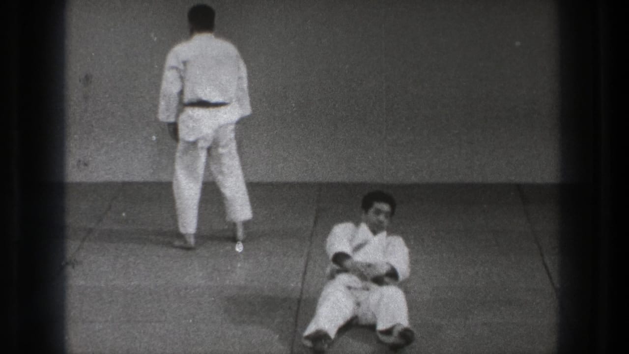 One adult in a white martial arts uniform stands facing the wall while another sits on the mat in this 1971 Tokyo practice footage.