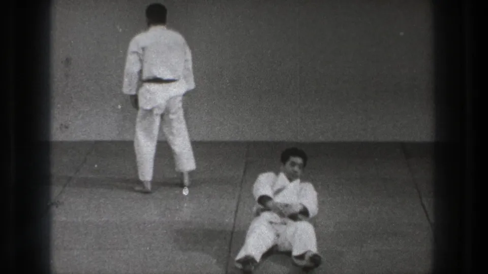 One adult in a white martial arts uniform stands facing the wall while another sits on the mat in this 1971 Tokyo practice footage.