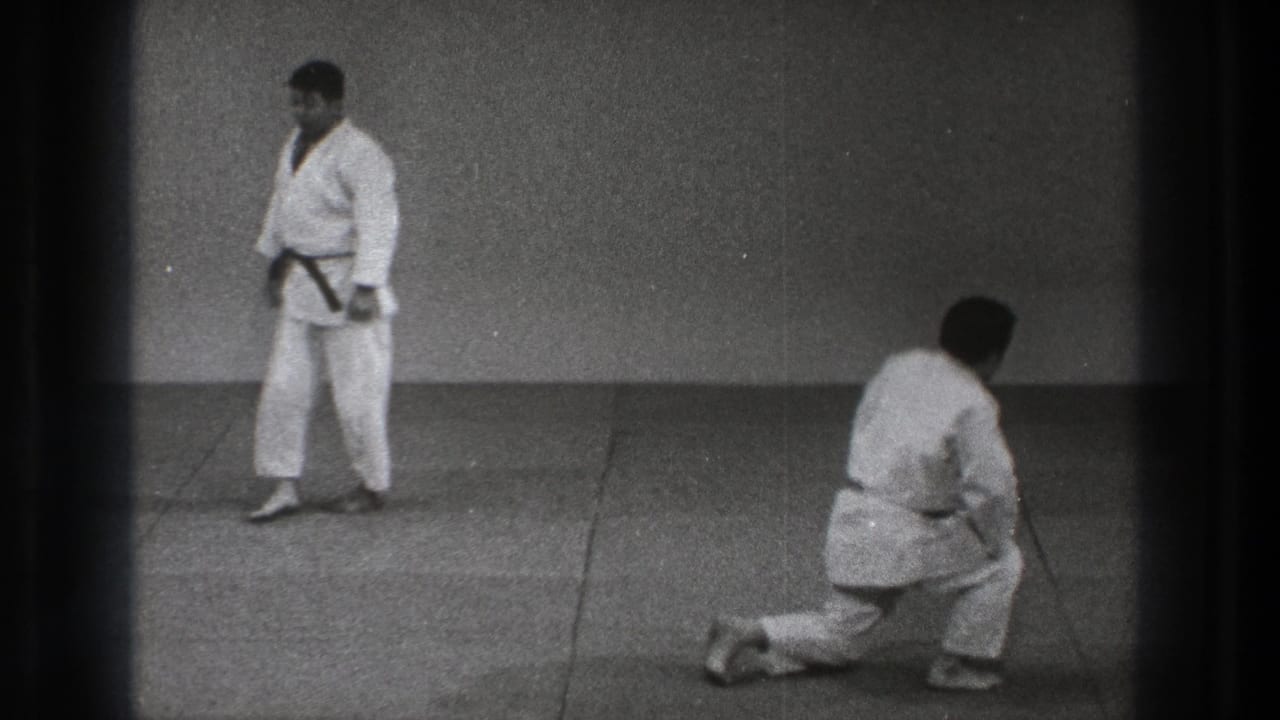 Two men in karate gi on a mat, with one kneeling and one standing.