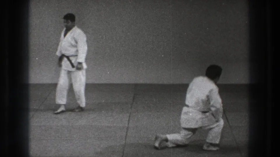 1971 Tokyo karate practice frame showing one man standing in a white gi while another kneels on a mat nearby.
