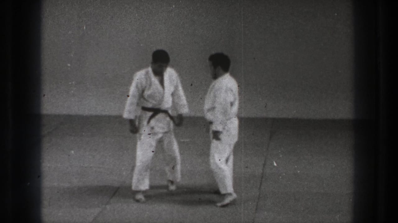 Two men in white gi stand close during a 1971 Tokyo karate practice sequence on a plain indoor mat.