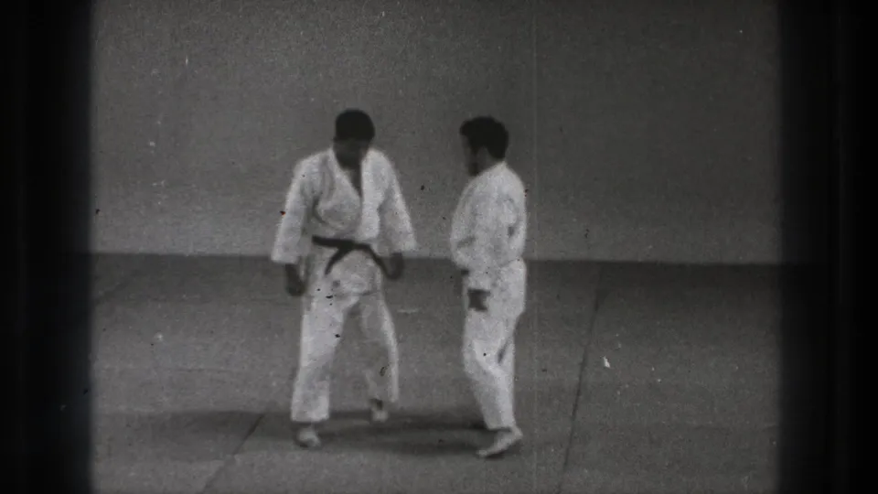 Two men in white gi stand close during a 1971 Tokyo karate practice sequence on a plain indoor mat.