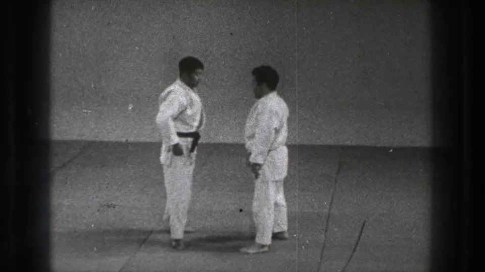 In Tokyo 1971 footage, two adult martial artists stand facing each other on a plain mat, one in a black belt and one in a lighter belt.