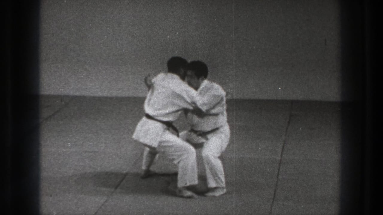 The practitioners lower into a close grappling position during the Uki Waza demonstration on a simple indoor mat.