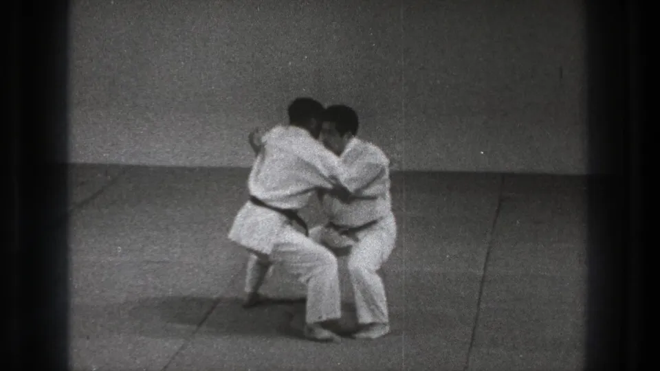 The practitioners lower into a close grappling position during the Uki Waza demonstration on a simple indoor mat.