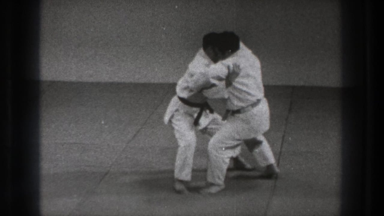 Two men in white gi practice a close grappling hold on a padded floor.