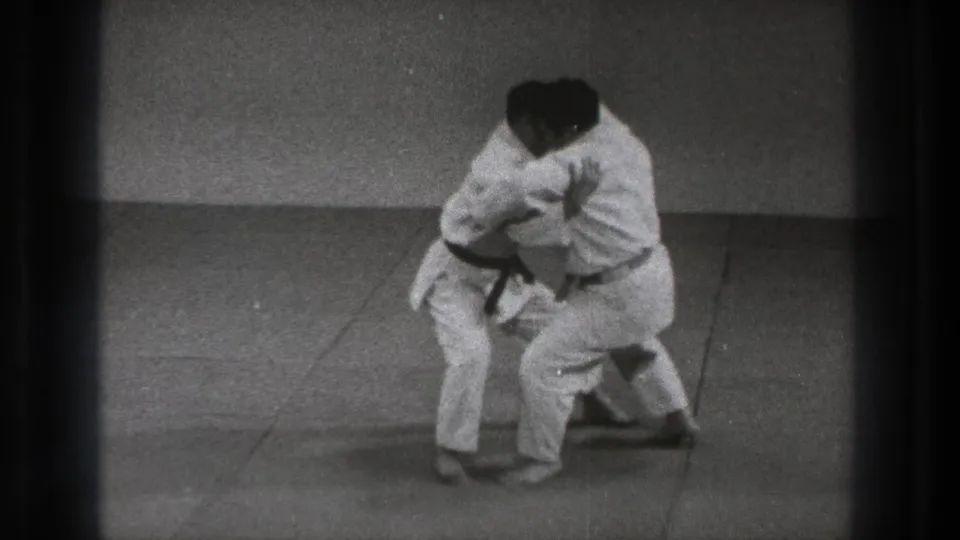 In 1971 Tokyo footage, two adult men in white gi work through a close martial arts hold on a plain padded floor.