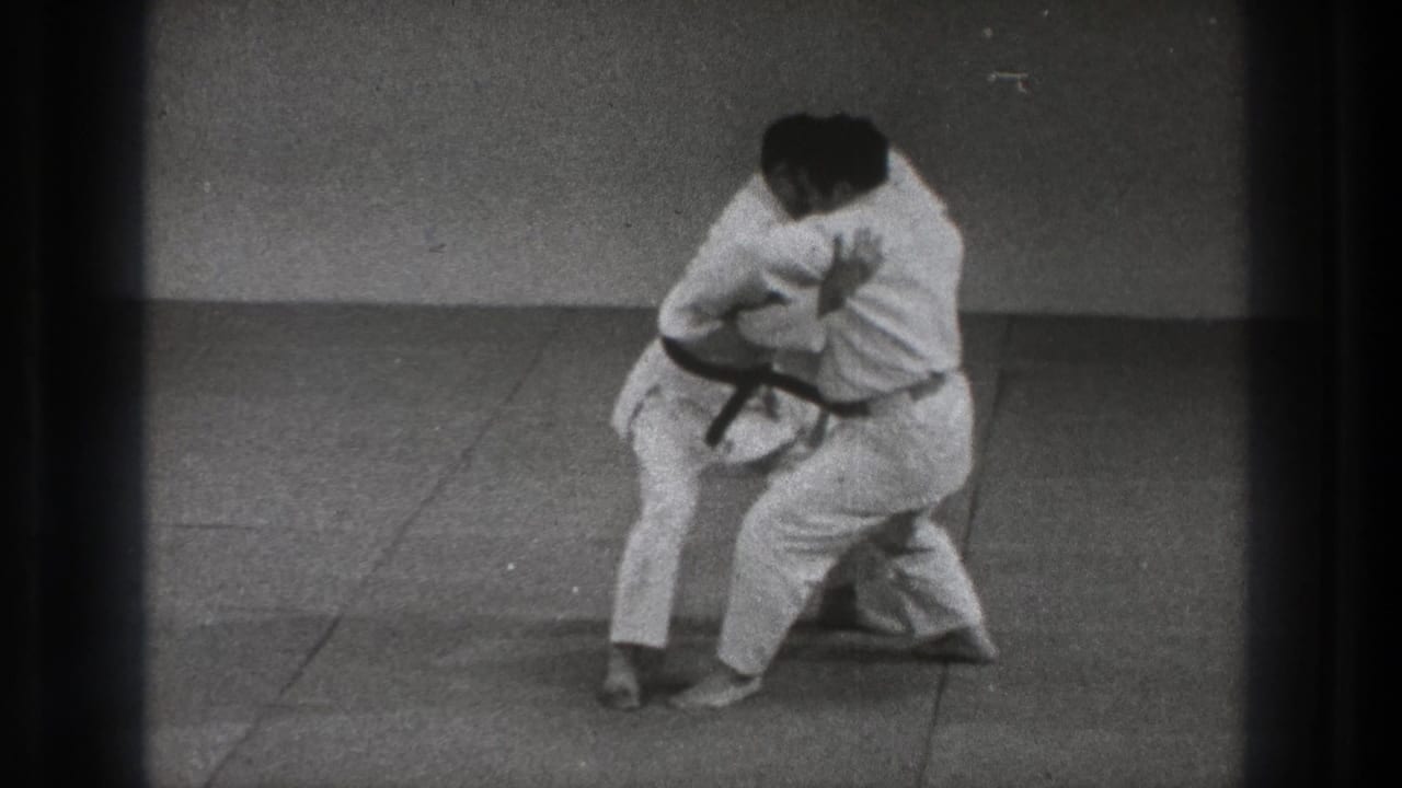 Two adult martial arts practitioners in white gi maintain a close hold during Tokyo training footage from 1971.