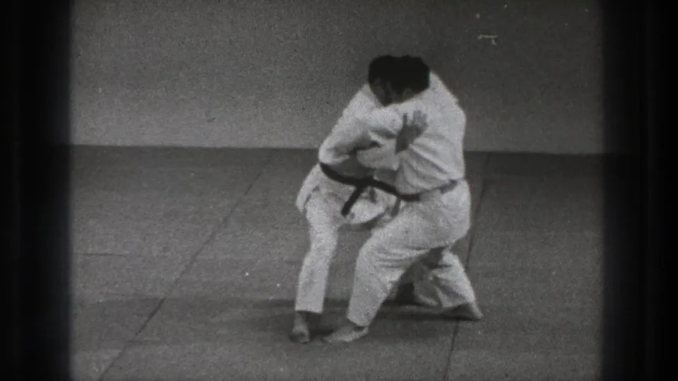Two adult martial arts practitioners in white gi maintain a close hold during Tokyo training footage from 1971.