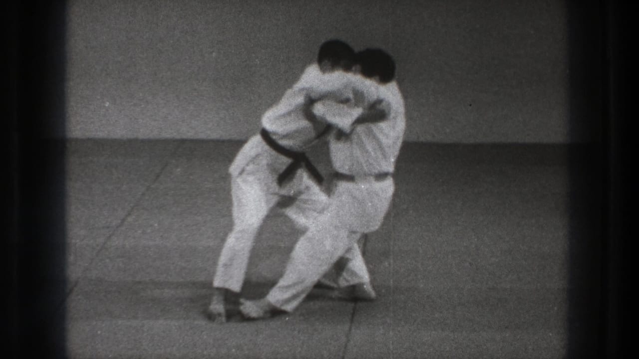 Two men in white martial arts uniforms grapple on a mat near a plain wall.