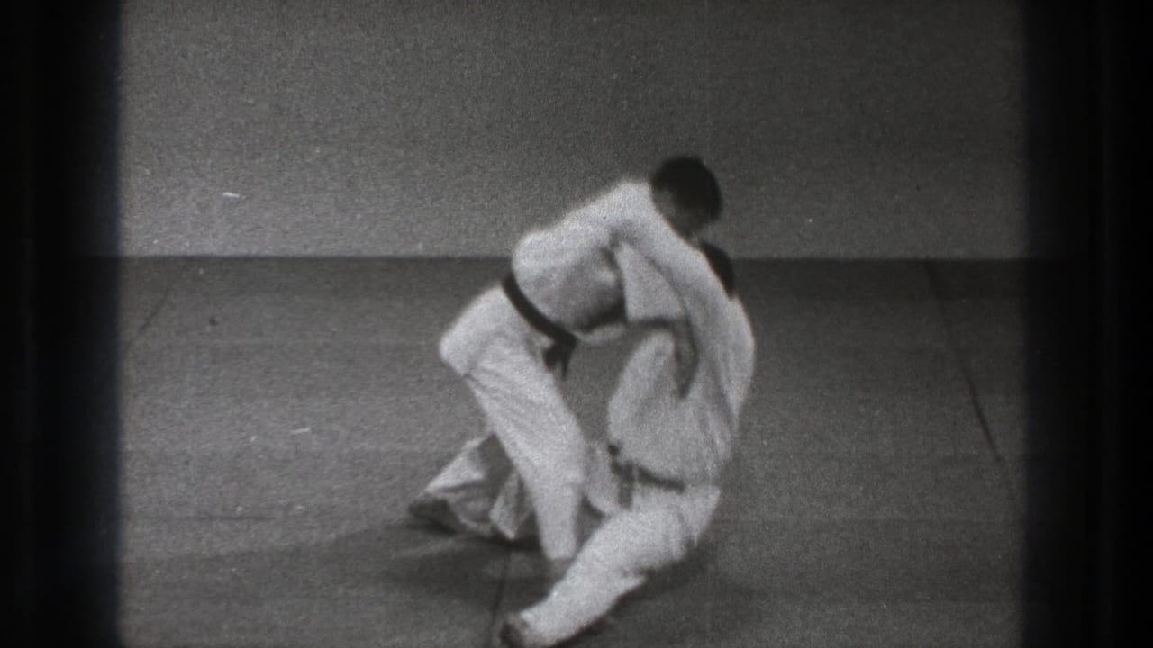 A martial arts throw reaches the fall, with one man bending forward and the other dropping onto the mat in the 1971 Tokyo training footage.