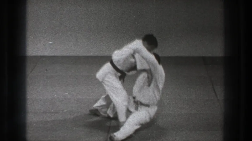 A martial arts throw reaches the fall, with one man bending forward and the other dropping onto the mat in the 1971 Tokyo training footage.