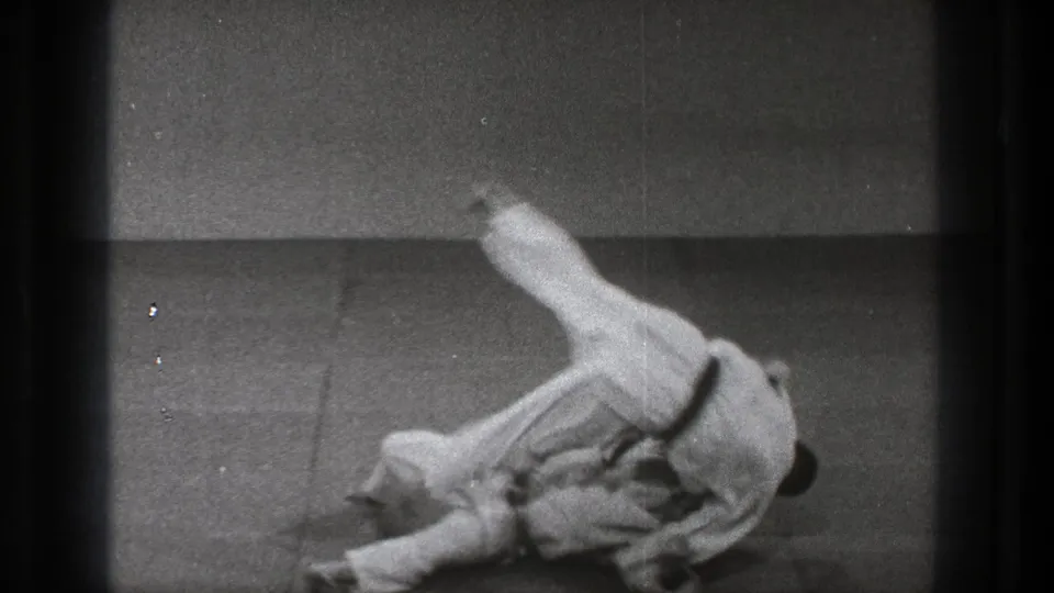 Two adult martial artists in white judogi roll on a mat in a simple Tokyo training-room scene from 1971 footage.
