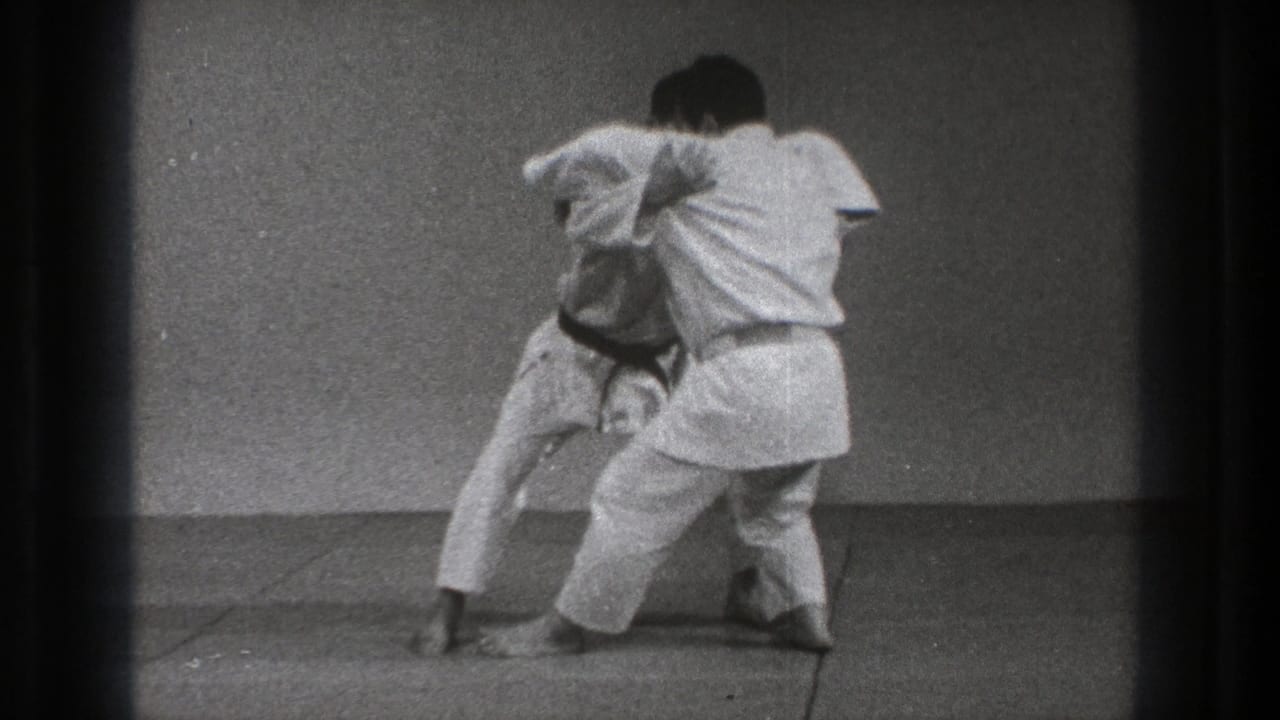 Two adult judo practitioners in white uniforms reset in a standing grip during the supported 1971 Tokyo training sequence.