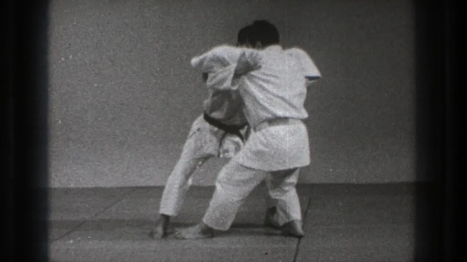 Two adult judo practitioners in white uniforms reset in a standing grip during the supported 1971 Tokyo training sequence.