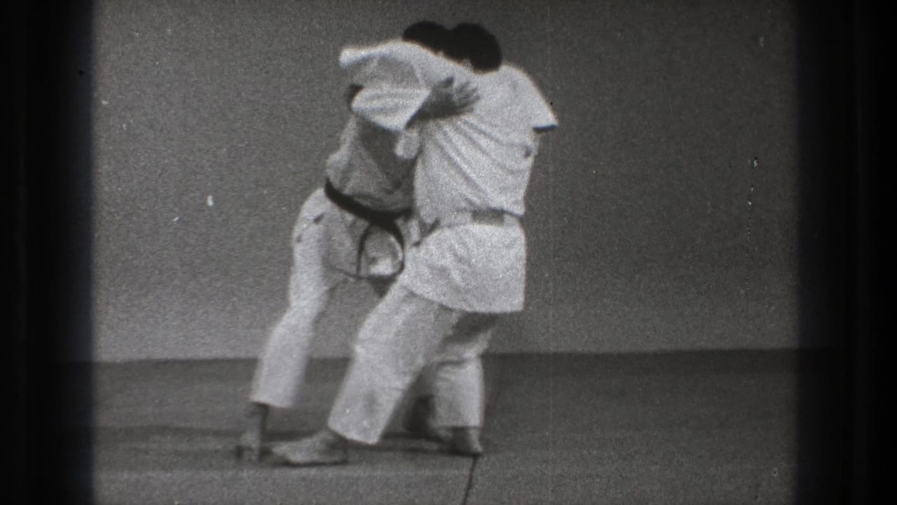 Two martial artists in white gi grapple on a mat during a throw setup.