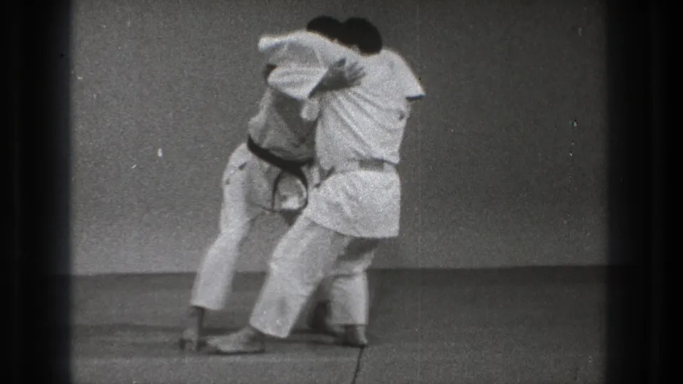 In supported 1971 Tokyo footage, two adult martial artists in white gi make close contact on a plain indoor mat as a throw begins.