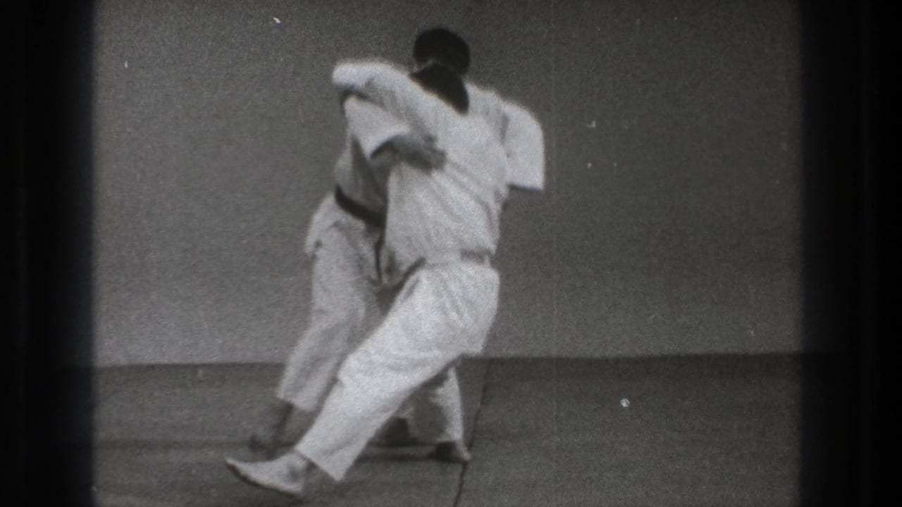 The throw develops as one gi-clad practitioner pivots sideways with the other partly obscured against a simple indoor practice space.