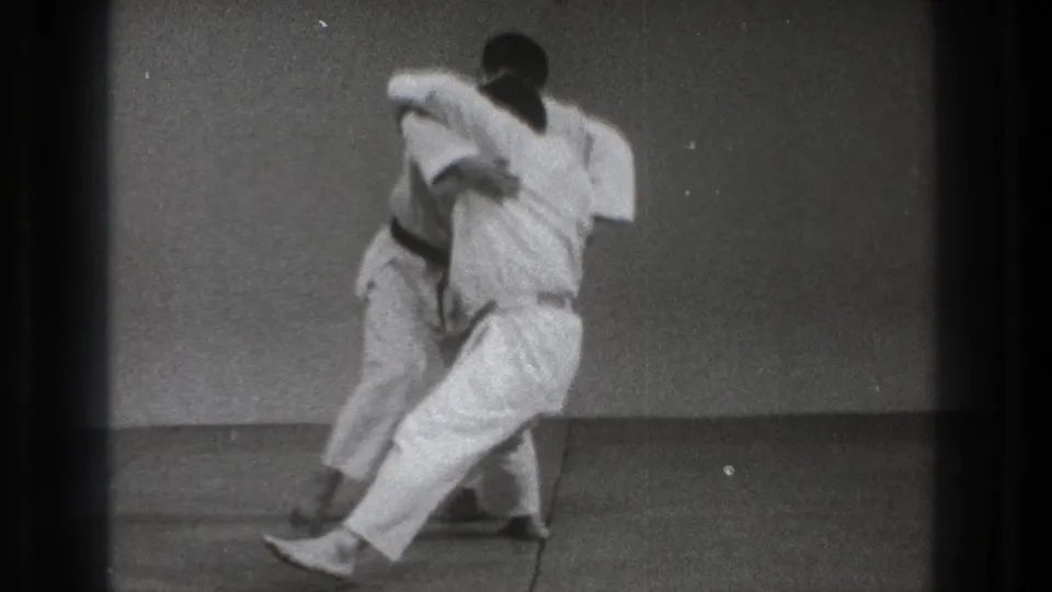 The throw develops as one gi-clad practitioner pivots sideways with the other partly obscured against a simple indoor practice space.