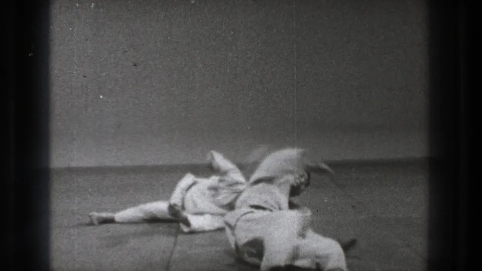 After the throw, one practitioner in a white gi is down on the mat, with the other partly visible at the edge of the frame.